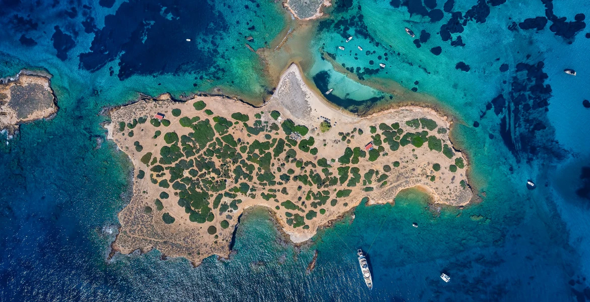 Top-down drone shot of Idrousa with sparse greenery and turquoise water.
