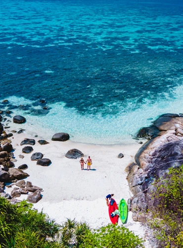 Aerial view of a couple walking on a white sand beach with kayaks and turquoise sea.