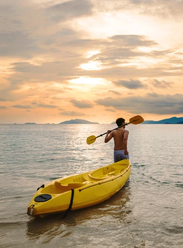 Man carrying a paddle, preparing to kayak into the sea at sunset.